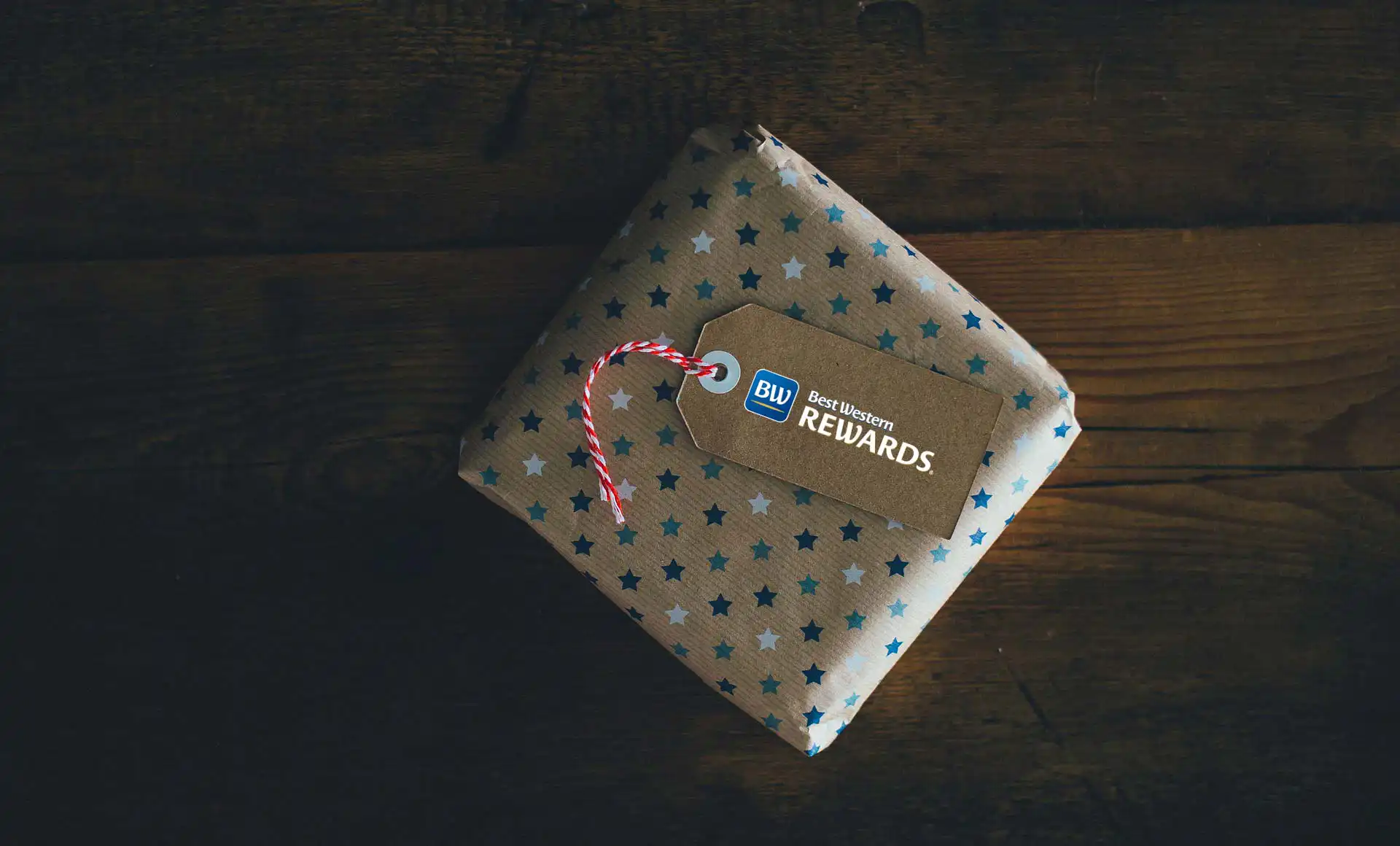 The image shows a gift package with brown paper and blue and light blue stars. On top of the package is a brown tag with a red and white ribbon, and the BW Rewards logo is printed on it.