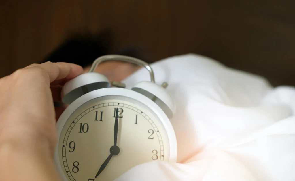 The image shows a vintage white alarm clock resting on a bed's duvet. A man's hand rests beside it, touching the alarm clock. Behind it, his head can be seen, lying under the covers.