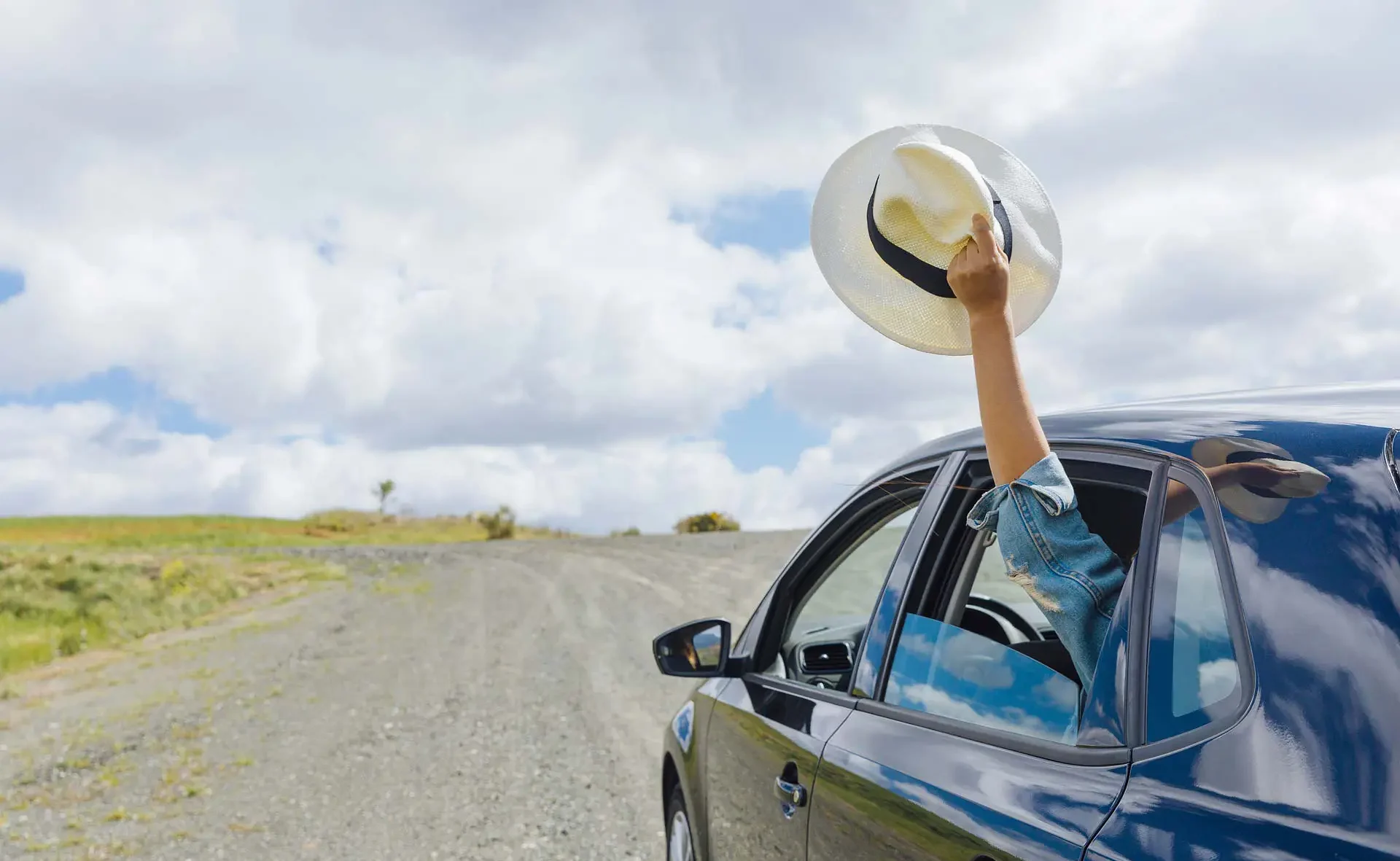 The image shows a girl's arm sticking out of the back window of a blue car. She's holding a straw hat in her hands and holding it up. Ahead, the road is a dirt road surrounded by greenery.